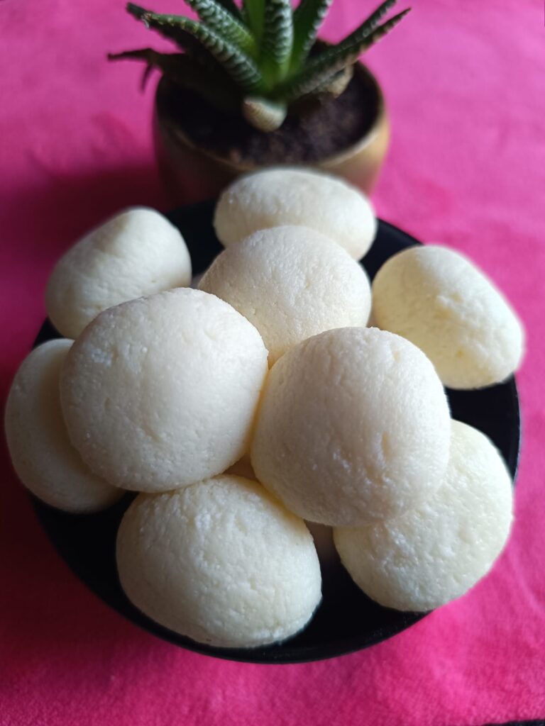 Bengali rasgullas are kept in a bowl on a pink color towel and a small plant is there near the bowl for decoration